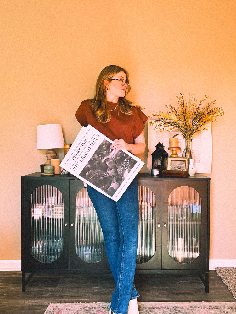 Graphic designer standing against a wall, holding up the Pigeon Post branding newspaper.